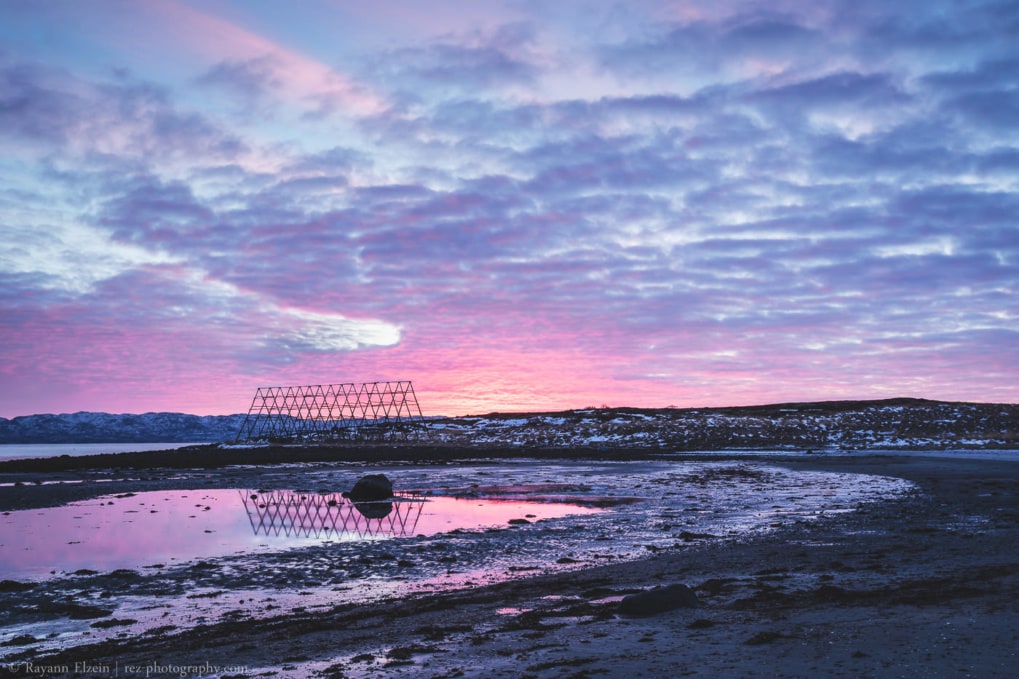Fish drying rack under bright pink sky reflecting in a tidal pool in Nesseby, Norway