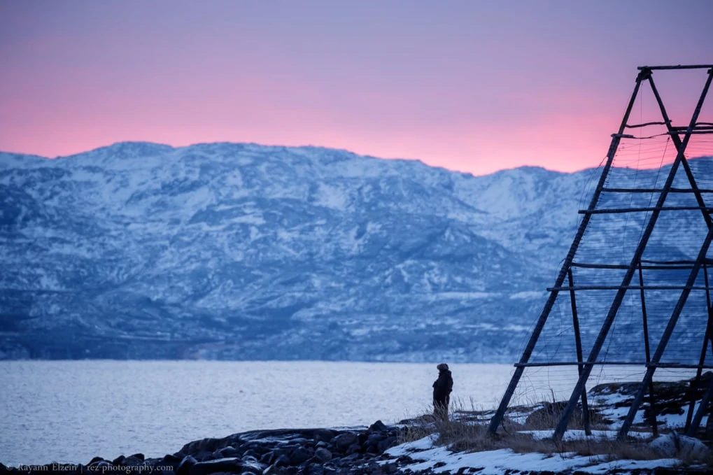 Man standing on the shore of the fjord, with the mountains and bright pink sky in the background